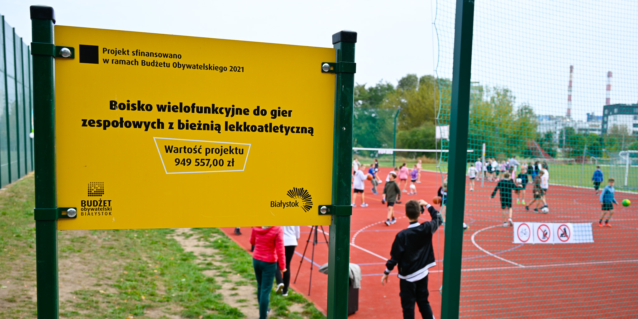 Zdjęcie tablicy z informacjami o boisku wielofunkcyjnym. W tle uczniowie grają w gry.