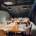Uczniowie siedzą przy stolikach. Robią eksperymenty. Są ubrani w białe fartuchy i okulary ochronne.