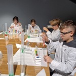 Uczniowie siedzą przy stolikach. Robią eksperymenty. Są ubrani w białe fartuchy i okulary ochronne.
