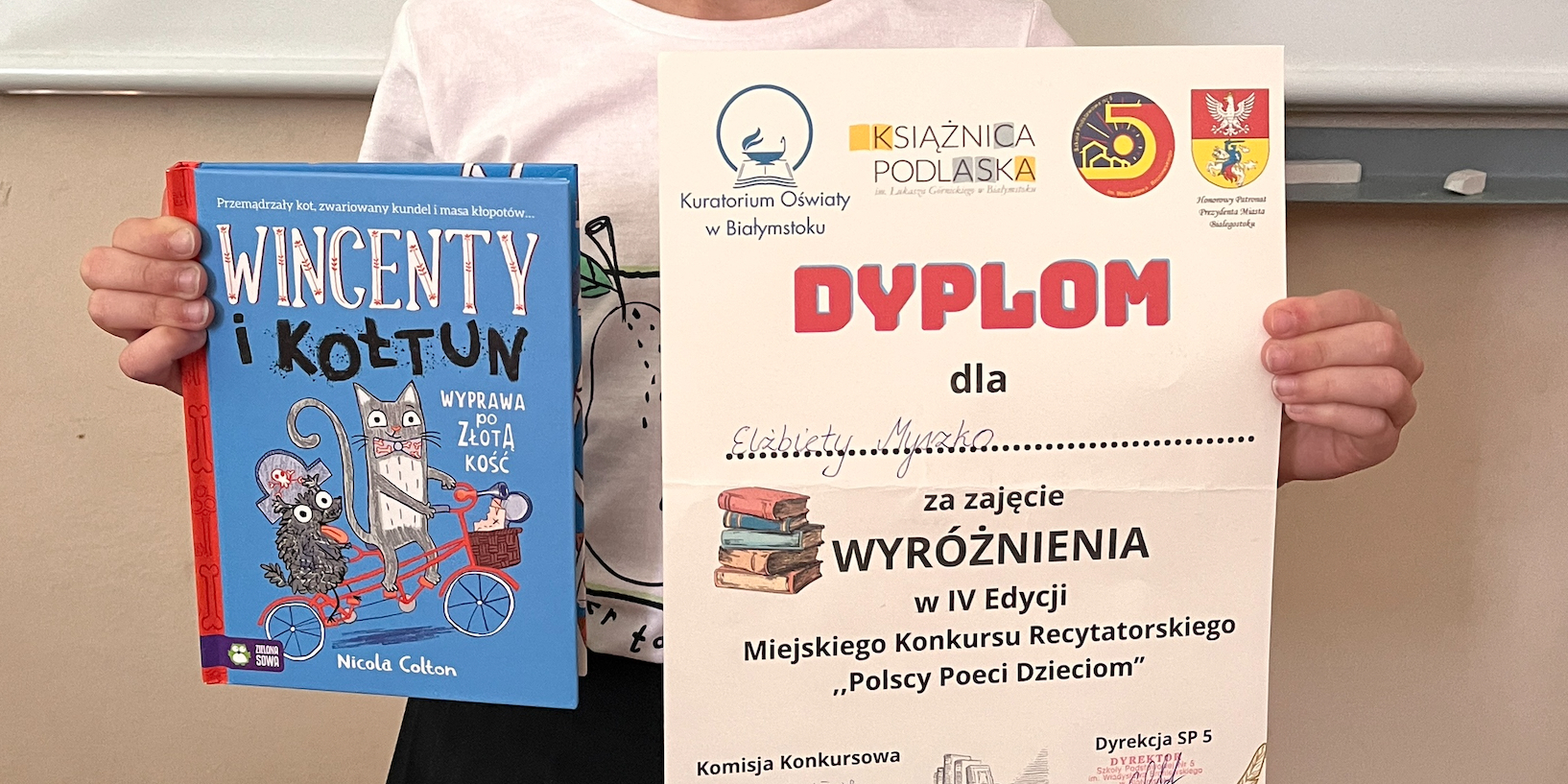 Uczennica pozuje do zdjęcia z dyplomem i nagrodą ksiązkową.