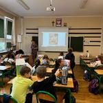 Ucznniowie siedzą w ławkach w sali. Nauczyciel czyta im książkę. Wyświetla treści z rzutnika na tablicę