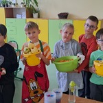 Uczniowie pozują do zdjęcia. Są ubrani w fartuszki kuchenne. Trzymają w rękach miski z obranymi owocami i warzywami