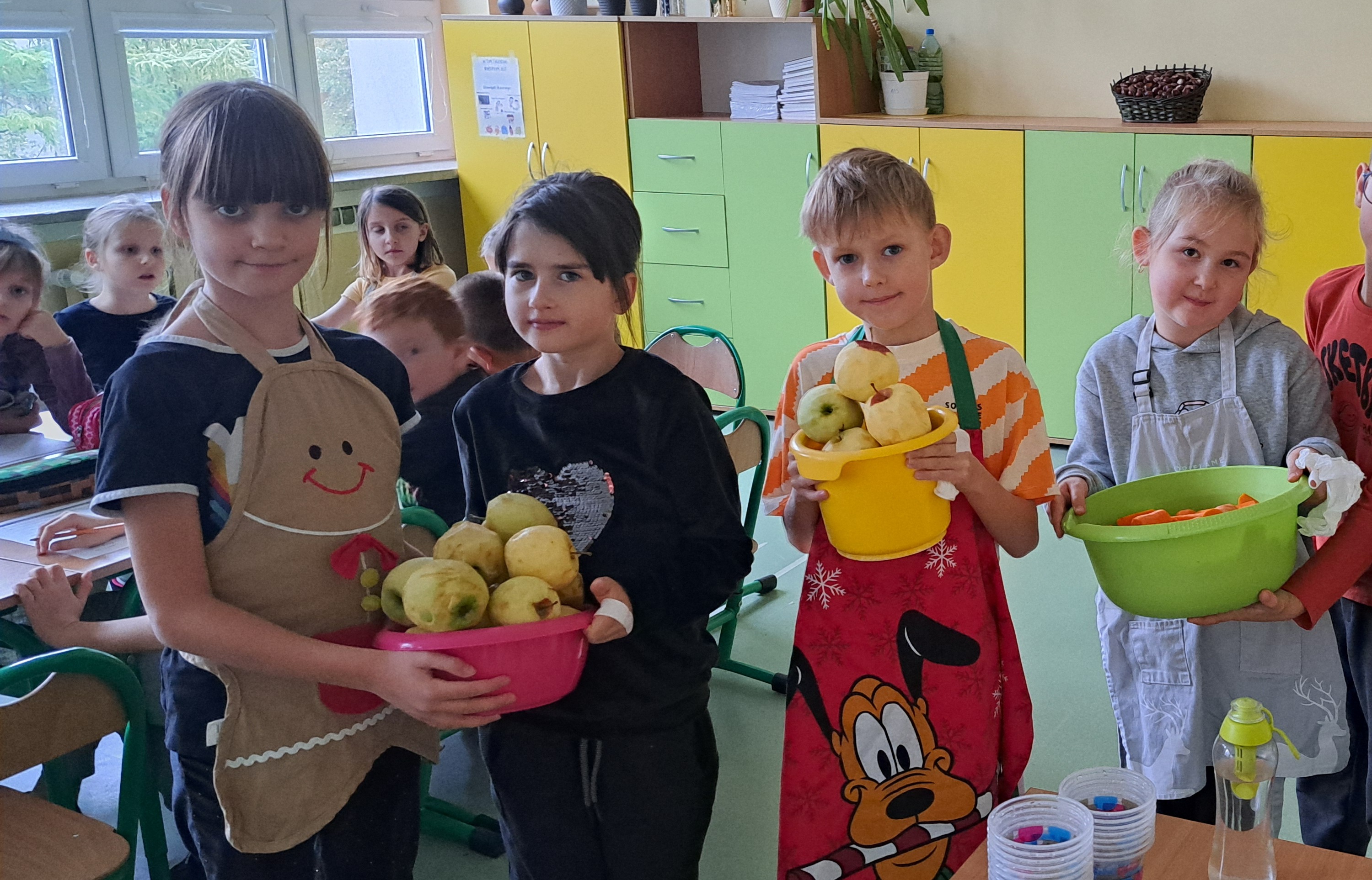 Uczniowie pozują do zdjęcia. Są ubrani w fartuszki kuchenne. Trzymają w rękach miski z obranymi owocami i warzywami..jpg