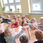 Uczniowie siedzą przy wspólnym stole. Podnoszą do góry kubeczki z sokiem