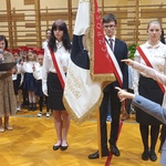 Trzech uczniów z pocztem sztandarowym stoi na środku. Pierwszoklasiści z wyciągniętymi przed siebie rękoma ślubują.
