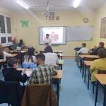 Nauczyciel stoi na środku sali_ uczniowie z rodzicami siedzą w ławkach.  Nauczyciel dyktuje treśc dyktanda
