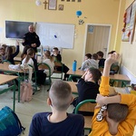 Uczniowie biorą udział w lekcji. Siedzą w ławkach. Jeden z uczniów podnosi rękę.
