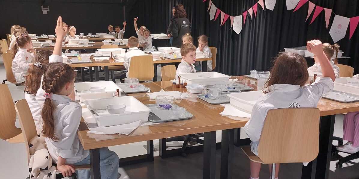 Uczniowie siedzą przy stołach. Ubrani są w białe kitle. Podnoszą ręce do góry - biorą udział w zajęciach..jpg