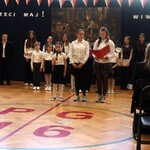 Uczennice na uroczystej akademii. Prezentują swoją rolę. Za nimi stoją inni uczestnicy akademii