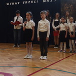 Uczniowie na uroczystej akademii. Prezentują swoje role. Za mini stoją inni uczestnicy akademii.