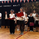 Uczennice na uroczystej akademii. Prezentują swoją rolę. Za nimi stoją inni uczestnicy akademii.