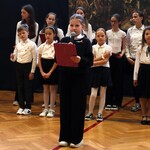 Uczennica na uroczystej akademii. Prezentuje swoją rolę. Za nią stoją inni uczestnicy akademii.