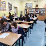 Uczniowie z rodzicami siedzą w ławkach. Piszą dyktando.