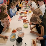 Ucznoiwie stoją dookoła stołu. Na stole na talerzach znajdują się składniki na pizzę. Uczniowie wybierają składniki