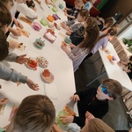 Ucznoiwie stoją dookoła prostokątnego stołu - szykują pizzę. Na stole na talerzach znajdują się składniki na pizzę