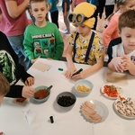 Uczniowie stoją dookoła stołu. Na stole na talerzach znajdują się składniki na pizzę. Uczniowie wybierają składniki