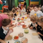 Ucznoiwie stoją dookoła prostokątnego stołu. Na stole na talerzach znajdują się składniki na pizzę