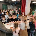 Uczniowie stoją dookoła prostokątnego stołu. Na stole na talerzach znajdują się składniki na pizzę W tle stoją uczniowie