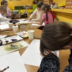 Dzieci siedzą przy stołach w klasie i rysują na białych kartkach. Na stolikach leżą farby, pędzle, kubeczki i przykładowe ilustracje. W tle widać żółte ściany, rośliny i półki z materiałami plastycznymi.