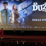 W sali kinowej trwa specjalny pokaz filmu „Za duży na bajki 2”. Na dużym ekranie wyświetlany jest plakat promocyjny z tytułem i postaciami z filmu. Przed ekranem siedzą dwie osoby na fotelach, a jedna osoba stoi i prowadzi wydarzenie. Na widowni widać publiczność zajmującą czerwone fotele.