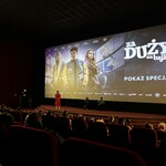 W sali kinowej trwa specjalny pokaz filmu „Za duży na bajki 2”. Na dużym ekranie wyświetlany jest plakat promocyjny z tytułem i postaciami z filmu. Przed ekranem siedzą dwie osoby na fotelach, a jedna osoba stoi i prowadzi wydarzenie. Na widowni widać publiczność zajmującą czerwone fotele.