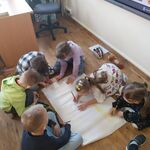 Kilka dzieci siedzi na podłodze w sali i rysuje na dużym arkuszu papieru_ otoczonym przyborami plastycznymi