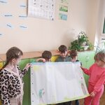 Grupa dzieci stoi w sali przy zielonych szafkach i trzyma duży arkusz papieru z rysunkami