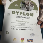 Osoba trzyma dyplom z wyróżnieniem w Wojewódzkim Konkursie Plastycznym S.O.S dla bezdomnych kotów!. Na dyplomie znajduje się zdjęcie kota oraz zielone elementy graficzne w formie łapek. Widoczny jest tekst z imieniem i nazwiskiem nagrodzonej osoby, nazwą szkoły oraz logotypy organizatorów. Na dole znajduje się data: Białystok, 17.02.2026 r