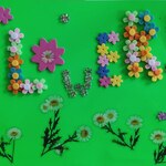 Zielone tło z kolorowym napisem FLOWERS ułożonym z małych piankowych i plastikowych kwiatków. Pod napisem znajduje się rząd białych stokrotek z zielonymi łodygami. Całość wygląda jak kolorowa, ręcznie wykonana dekoracja plastyczna.