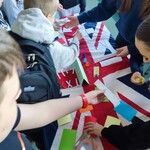 Uczniowie stoją wokół stołu przykrytego flagą Wielkiej Brytanii i wspólnie pracują z kolorowymi kartonikami, kartkami oraz drobnymi elementami podczas aktywności edukacyjnej.