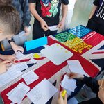 Uczniowie stoją wokół stołu przykrytego flagą Wielkiej Brytanii i pracują z kartami z tekstem oraz kolorowymi elementami.