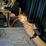 Zbliżenie na doświadczenie z iskrzeniem – dłonie osoby trzymają przewody, z których wydobywają się iskry, a uczniowie pracują przy stanowiskach w tle.