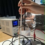Zbliżenie na doświadczenie z elektrodami zanurzonymi w cieczy podłączonymi do zasilacza laboratoryjnego.