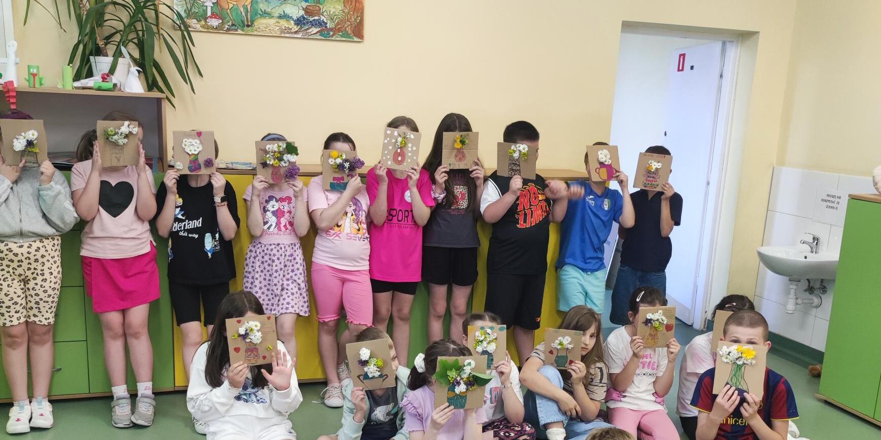 Grupa dzieci w klasie trzyma przed sobą kolorowe kartonowe prace plastyczne z kwiatami