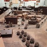 Makieta miasteczka wykonana z czekolady przedstawiająca budynki, ulice i drzewa; miniaturowa zabudowa miejska z blokami i domami otoczona zielonymi terenami i chodnikami, oglądana przez zwiedzających w przestrzeni wystawowej.